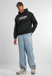 Felpa nera in cotone con tasca frontale e testo bianco. Abbinata a jeans baggy azzurri e scarpe da ginnastica grigie. Indossata con un cappellino nero.