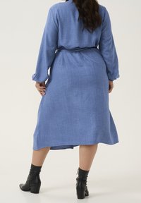 Blauwe jurk met lange mouwen in een gestructureerde stof, met een ceintuur rond de taille, midi-lengte en zijsplitten, gecombineerd met zwarte enkellaarsjes.