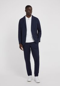 Jachetă de costum bleumarin cu două butoane și un buzunar, asociată cu un tricou polo alb. Pantaloni bleumarin slim-fit și adidași albi.