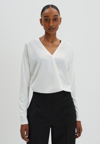 Haut blanc à manches longues avec un col en V et une coupe décontractée, associé à un pantalon noir ajusté, doté d'un tissu lisse et d'un design simple.