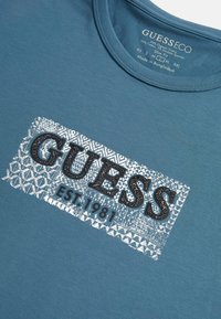 Tyrkysové bavlněné tričko s texturovaným černým logem "GUESS" a stříbrným geometrickým vzorem na pozadí, pod kterým je uvedeno "EST. 1981".