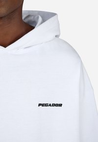 Λευκή κουκούλα από μαλακό ύφασμα με μια έκφραση μπροστινής τσέπης καγκουρό. Διαθέτει μαύρο κεντημένο κείμενο "PEGADOR" στο αριστερό στήθος.