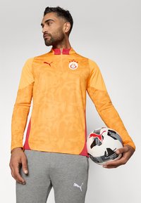 Pomarańczowa i żółta bluza treningowa z długim rękawem, w wzory, z czerwonymi akcentami i kołnierzem na pół-zamek. Szare dresy z białym logo; trzymająca piłkę nożną.