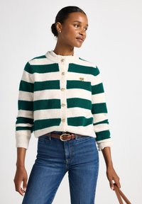 Grøn- og hvidstribet cardigan med rund halsudskæring, knaplukning og hjerteformet accent. Båret over blå jeans og et brunt bælte.