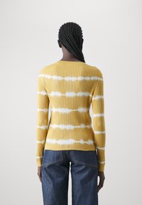 Żółty sweter prążkowany z poziomym białym wzorem tie-dye, z okrągłym dekoltem i długimi rękawami, w zestawieniu z niebieskimi dżinsami o podwyższonym stanie.