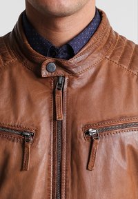 Giacca di pelle marrone con colletto alto, dotata di due tasche sul petto con zip, cuciture a vista e una texture liscia.