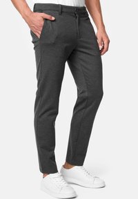 Homme portant un pantalon slim gris foncé avec bouton et fermeture éclair, des baskets blanches, posant avec une main dans la poche sur fond blanc.