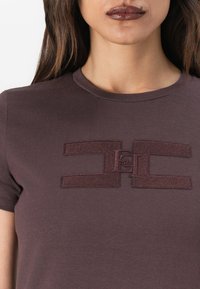 T-shirt à manches courtes et col rond en mauve foncé avec un logo texturé en relief au centre, représentant deux formes entrelacées.