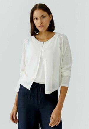 Vrouw met schouderlang bruin haar, gekleed in een witte gebreide cardigan met knopen over een witte top en donkerblauwe broek, staand tegen een effen achtergrond.