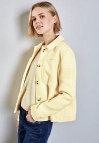 Gul rutig jacka, texturerat tyg, knappframsdesign, enfaldig bröstficka. Parat med en beige tröja och mörka jeans.