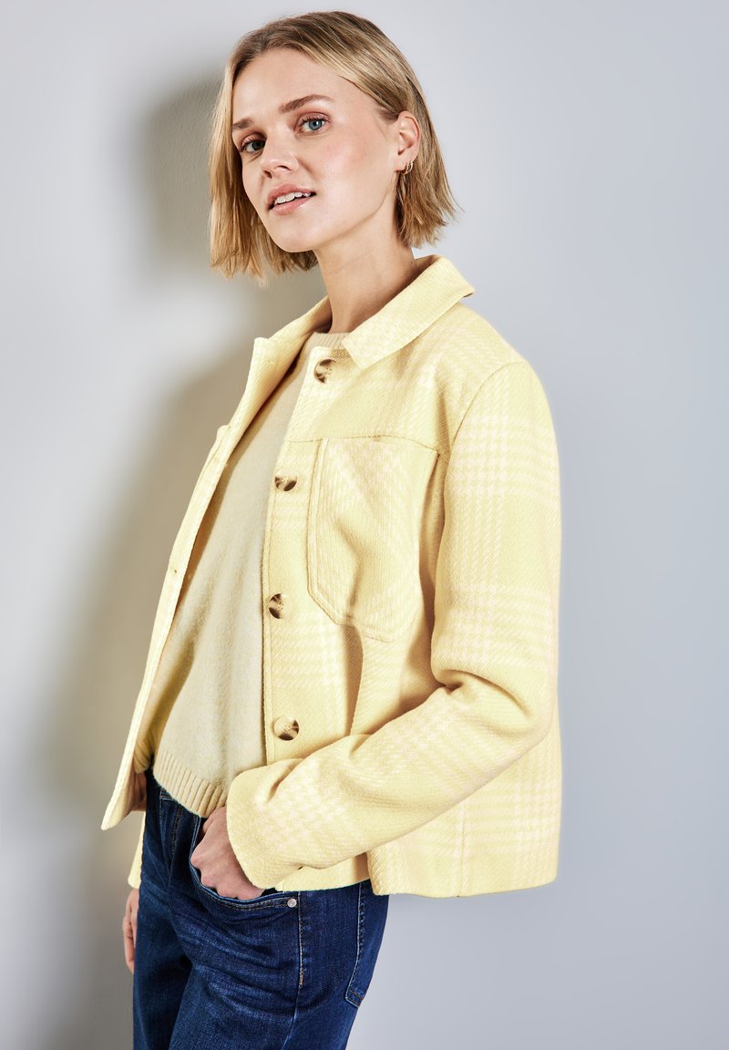 Gul rutig jacka, texturerat tyg, knappframsdesign, enfaldig bröstficka. Parat med en beige tröja och mörka jeans.