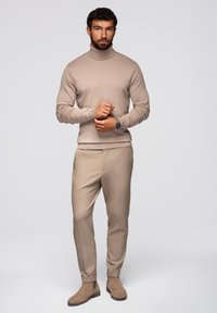 Maglione beige con collo alto e pantaloni sartoriali coordinati, caratterizzati da una texture liscia, abbinati a stivaletti beige. Design minimalista.