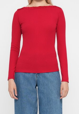 Nainen yllään tiukka punainen pitkähihaisen puseron pitsireunuksella kaula-aukossa ja väljät siniset farkut, seisoo yksinkertaista taustaa vasten.