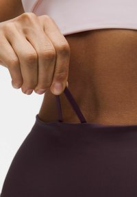 Abbigliamento attivo viola aderente con una texture liscia; una mano tira una cinghia coordinata dall'elastico della vita, su uno sfondo neutro.