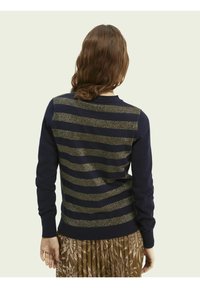 Suéter azul marino con franjas horizontales doradas brillantes, puños acanalados y cuello redondo. Llevado sobre una falda plisada y estampada.