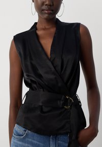 Black satin wrap top with a deep V-neck, slim belt with gold hardware, and sleeveless design. Paired with light blue denim.