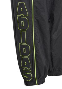 Un pantalon coupe-vent noir présente un texte "ADIDAS" en néon vert audacieux le long du côté et une bande contrastante vert citron. Des poignets élastiques aux chevilles.