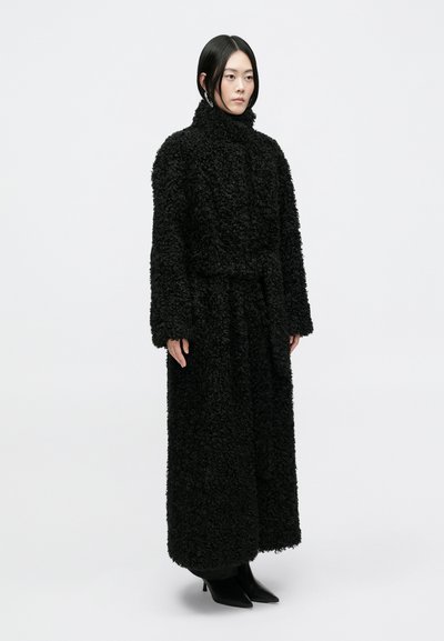 Casaco preto, fofinho, de pelagem longa com um colarinho alto, detalhe de cinto e mangas largas. Usado sobre um design até ao chão com textura suave.