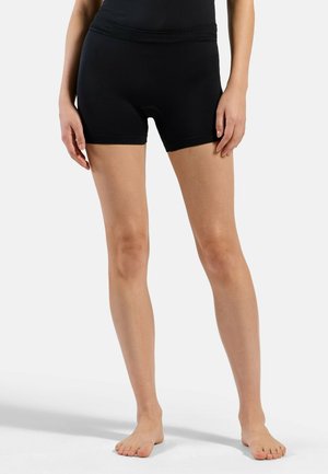 Person trägt schwarze, enganliegende Radhosen, steht barfuß vor einem schlichten hellen Hintergrund, Hände ruhen seitlich.