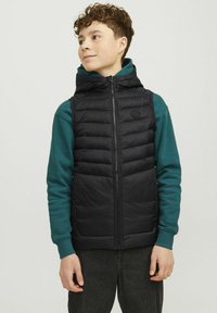 Crni puffa prsluk s visokim ovratnikom i patentnim zatvaračem, složen preko teal dugih rukava. Sadrži kviltanu teksturu i ramene šavne.