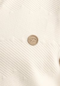 Roomkleurige, getextureerde stof met een geribbeld ontwerp en een cirkelvormig beige embleem. Het materiaal heeft een subtiele glans en verhoogde patronen.