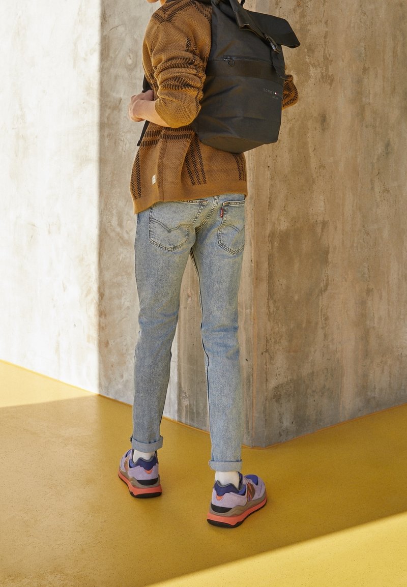 Pull en tricot marron avec texture à motifs, jeans slim bleu clair et baskets multicolores avec des accents violets, roses et gris. Sac à dos noir.