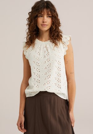 Kobieta o kręconych włosach, ubrana w białą ażurową bluzkę bez rękawów i brązową plisowaną spódnicę, stojąca na jednolitym tle.