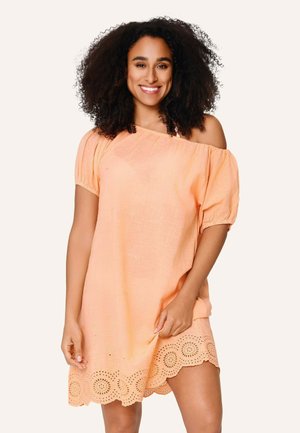 Mujer sonriente con cabello rizado, vestida con un vestido melocotón con hombros descubiertos y bajo de encaje calado, posando frente a un fondo claro y liso.