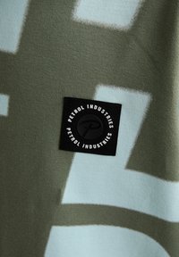 Oliemgroene stof met lichtblauwe geometrische patronen, voorzien van een zwart label met de tekst "Petrol Industries" in witte letters.