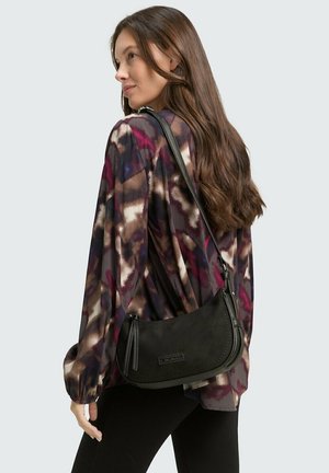 Schwarze Wildleder-Umhängetasche mit geschwungenem Design, Reißverschluss-Akzenten und einem schmalen, verstellbaren Riemen, die über einer gemusterten Bluse getragen wird.