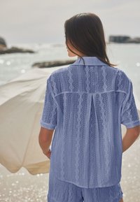 Donna con camicia azzurra a motivi e pantaloncini, vista di spalle, in piedi di fronte al mare con un ombrello beige sulla riva in una giornata nuvolosa.