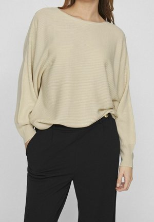 Kvinde iført en løs, beige ribstrikket sweater, der er stoppet ned i højtaljede sorte bukser, med den ene hånd i lommen og den anden afslappet ved siden.