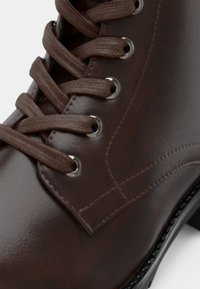 Brązowy skórzany botek za kostkę z okrągłym noskiem, sześcioma oczkami do sznurowania, teksturowaną powierzchnią i srebrnymi metalowymi oczkami, z płaską gumową podeszwą.