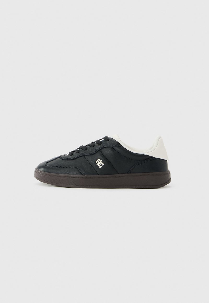Basket basse noire avec semelle marron, lacets noirs, col de talon blanc et petit logo blanc sur le côté.