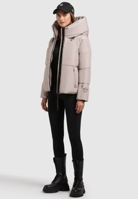 Helle beigefarbene Pufferjacke mit hohem Kragen, Reißverschluss und Seitentaschen. Kombiniert mit schwarzen Leggings und knielangen Schnürstiefeln.