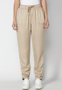 Pantalones jogger beige hechos de una tela ligera con una banda elástica en la cintura, dos bolsillos frontales y detalles fruncidos en el tobillo.
