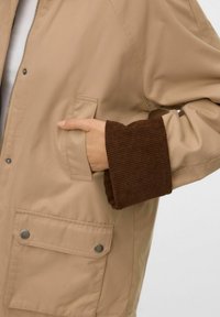 Chaqueta marrón con una cubierta suave, bolsillo de parche abotonado y detalles de puños acanalados en marrón. Cuenta con cierre de cremallera y un diseño elegante.