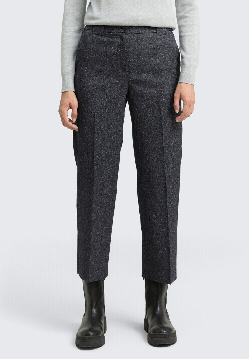 Personne portant un pantalon gris foncé ajusté, un haut à manches longues gris clair et des bottines noires, debout sur un fond uni.