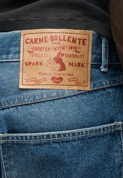 Svetlomodré denimové džínsy s hnedou koženou záplatou s červeným textom. Záplata obsahuje grafiku zajacov a dekoračné prvky.