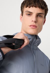 Lightweight, grey nylon jacket with a high collar, featuring a zip closure and black straps held in hand. Smooth texture, minimal branding.
