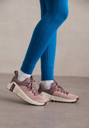 Persona con mallas azules, calcetines blancos y zapatillas deportivas malva con suelas blancas y gruesas, de pie sobre un suelo gris liso.