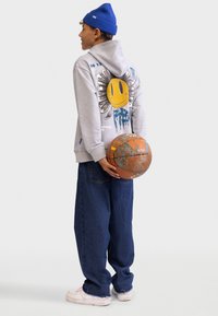 Felpa grigia con una stampa di smiley colorato e accenti blu, abbinata a jeans blu larghi e scarpe da ginnastica bianche, mentre tiene un pallone da basket consumato.