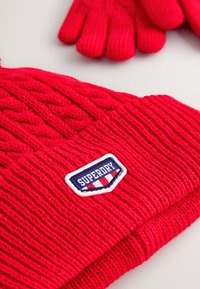 Rød strikket hue med ribbet design, der har et kabelstrikket mønster og en hvid lap med teksten "SUPERDRY." Inkluderer matchende røde handsker.