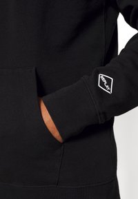 Sudadera con capucha negra de tela texturizada, con un bolsillo frontal, puños acanalados y un logo blanco "REPLAY" en la manga.