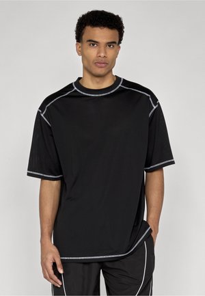 Młody mężczyzna ubrany w czarny, oversize'owy T-shirt z białymi przeszyciami i czarne spodnie z białym paskiem, stojący na jednolitym szarym tle.