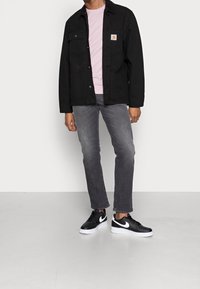 Chaqueta negra de lona con dos bolsillos delanteros, camiseta rosa debajo, jeans gris y zapatillas Nike en blanco y negro. Textura suave a lo largo.