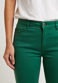Pantalon en mélange de coton vert à la texture lisse, doté d'une braguette zippée, d'une fermeture par bouton et de détails de poches avant.