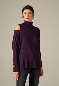 Maglione viola a collo alto con design spalle scoperte, polsini a coste e tessuto testurizzato. Indossato con jeans grigio scuro.