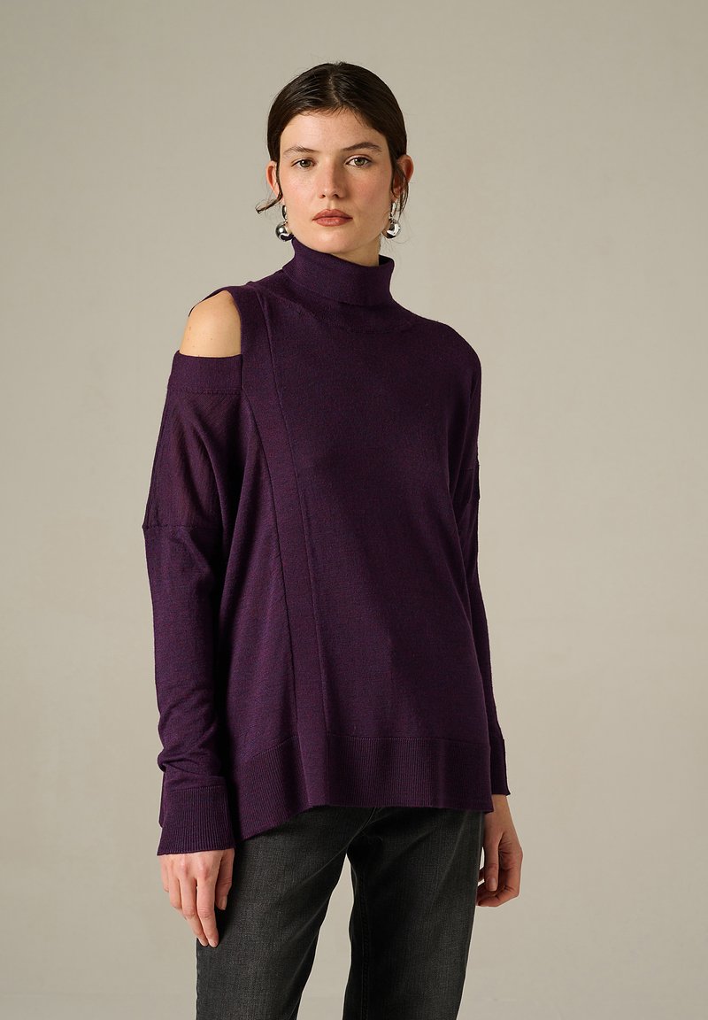 Maglione viola a collo alto con design spalle scoperte, polsini a coste e tessuto testurizzato. Indossato con jeans grigio scuro.