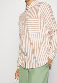 Camicia button-up a righe verticali pesca e bianche, con taschino sul petto e maniche lunghe. Abbinata a pantaloni verde chiaro.